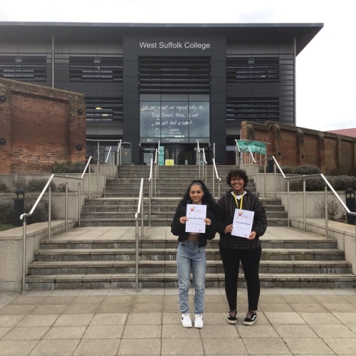 Photo of two people holding a certificate each in front of a West Suffolk College building