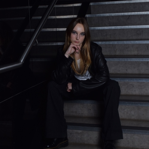 Person sitting on a staircase posing in a black leather jacket and pin stripped trousers