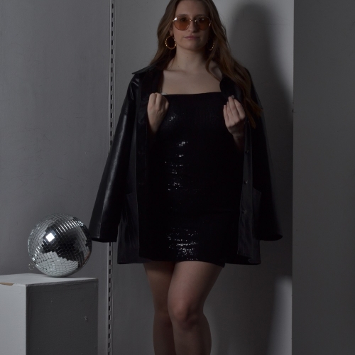 Person posing in a black dress, black leather jacket on her shoulders and beside her is a disco ball