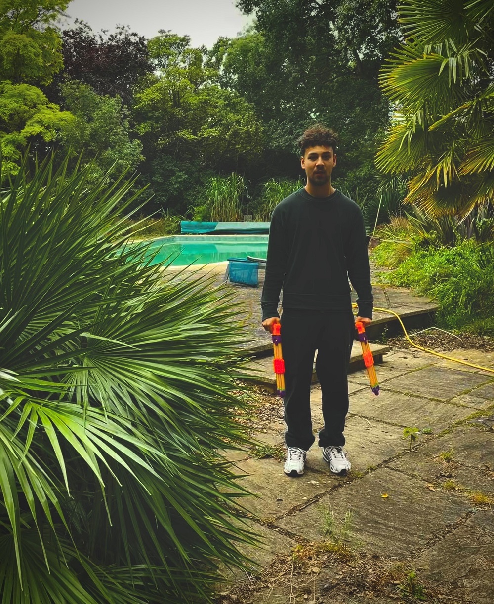 Ollie DeAlwis Music Artist - a boy in his teens surrounded by greenery holding two water canons